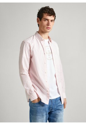 Camisa de manga larga a rayas en rosa claro y blanco, abotonada con un cuello, llevada sobre una camiseta blanca. Combinada con jeans de mezclilla azul claro.