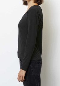 Kvinna som står i profil iklädd en svart långärmad tr öja och mörka jeans mot en enkel vit bakgrund.