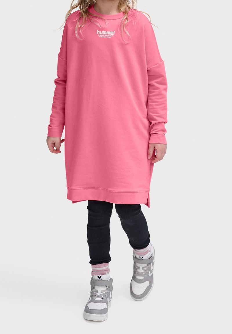 Enfant portant une longue robe sweat-shirt rose, des leggings noirs et des baskets montantes grises, marchant devant un fond clair uni.
