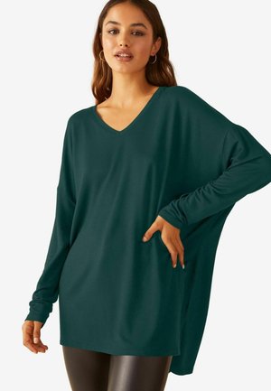 Langärmliges, übergroßes V-Ausschnitt-Oberteil aus tiefgrünem Stoff, das einen lockeren Schnitt und eine weiche Textur aufweist, getragen mit glänzenden schwarzen Leggings.