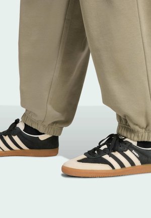 Černé a krémové sportovní boty s gumovými podrážkami, se třemi černými pruhy, z semišového a koženého materiálu, v kombinaci se šedými kalhotami.