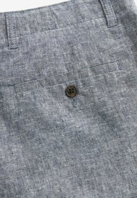 Gros plan sur un pantalon en tissu gris texturé avec une poche arrière, un bouton foncé et deux passants de ceinture à la taille.