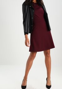 Robe bordeaux avec un corsage ajusté et une jupe évasée, associée à une veste en cuir noire. Des chaussures à talons hauts noires complètent la tenue.