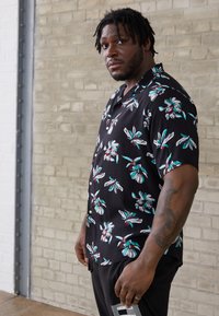 Short-sleeve black shirt with white and teal floral pattern, featuring buttons down the front and a relaxed fit. Worn with black pants.