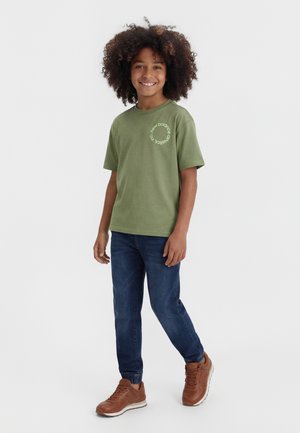 Smilende barn med krøllet hår iført grøn New Balance T-shirt, mørkeblå jeans og brune sneakers, stående foran en hvid baggrund.