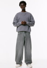Junger Mann mit dunklem, lockigem Haar, der einen lockeren grauen Sweatshirt, weite graue Jeans, weiße Turnschuhe und silberne Halsketten vor einem weißen Hintergrund trägt.
