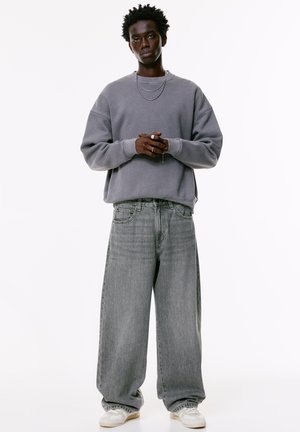 Young man with dark curly hair wearing a loose gray sweatshirt, wide gray jeans, white sneakers, and silver necklaces on a white background.