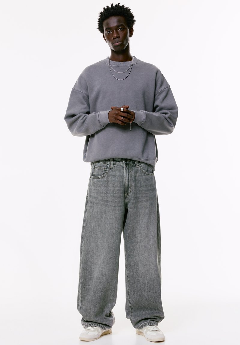 Junger Mann mit dunklem, lockigem Haar, der einen lockeren grauen Sweatshirt, weite graue Jeans, weiße Turnschuhe und silberne Halsketten vor einem weißen Hintergrund trägt.
