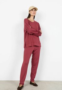 Set de îmbrăcăminte sportivă bordo format dintr-un top cu mâneci lungi și croială lejeră și pantaloni asortați, conici, completat cu o șapcă bej.