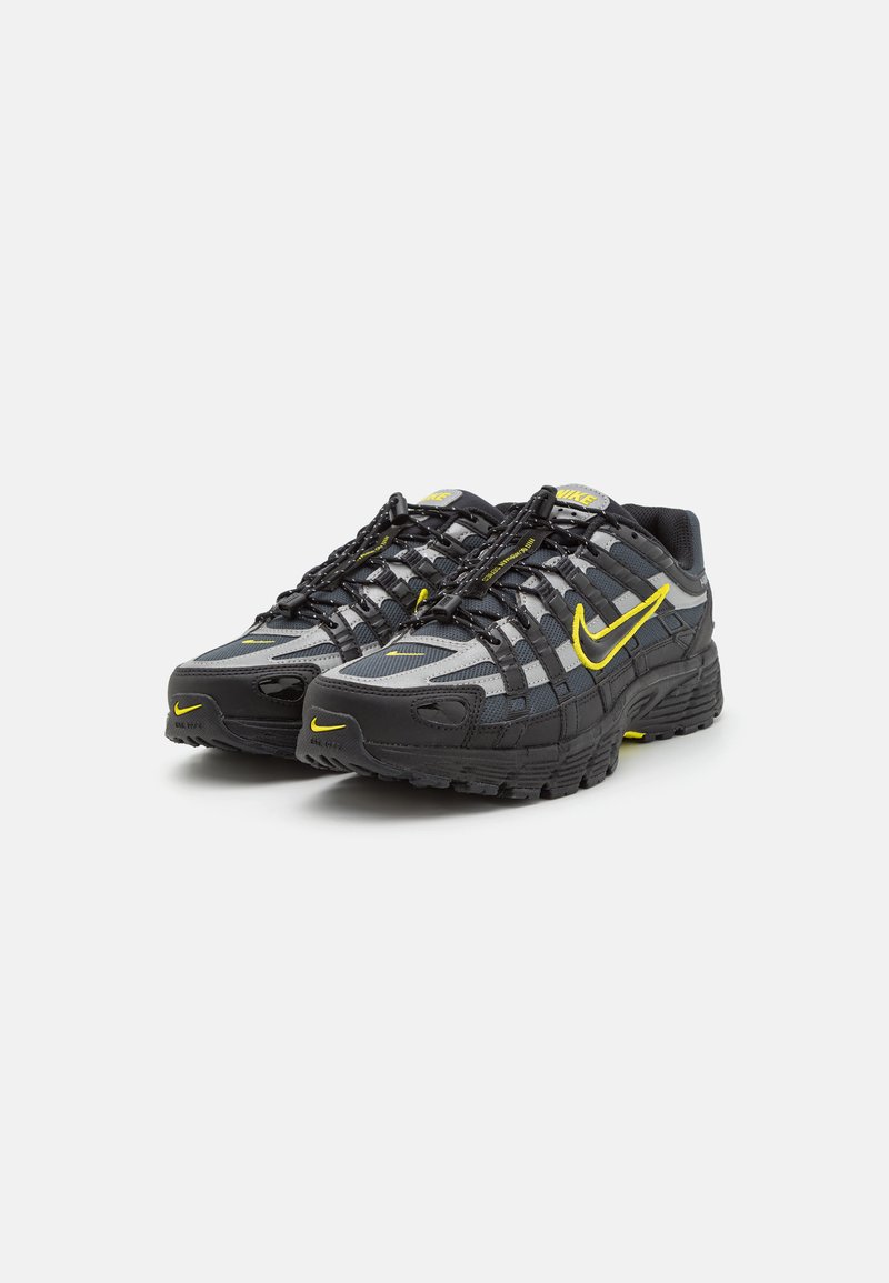 Zapatos deportivos en negro y gris con parte superior de malla, acentos en amarillo y un sistema de cordones único. La suela es texturizada para ofrecer tracción.