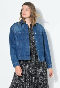 Denimjacka i mellanblått, med klassisk krage, två framfickor och knäppning. Bärs över en svart klänning med vita himlaföreställningar.