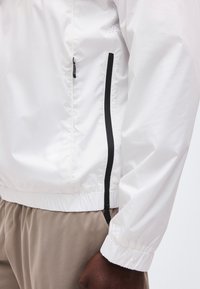 Κοντινό πλάνο ενός λευκού μπουφάν με μαύρη κάθετη τσέπη με φερμουάρ και ελαστική μανσέτα, φορεμένο με μπεζ παντελόνι.