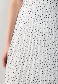Falda blanca plisada con lunares negros, que presenta una cintura ceñida. La tela es ligera, creando una apariencia texturizada.