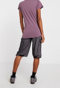 Violetti t-paita lyhyillä hihoilla, rento istuvuus. Mustat cargo-housut, joissa käänteet lahkeissa. Harmaat urheilukengät. Yksivärinen tausta.