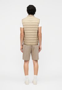 Colete acolchoado bege com gola alta, design em camadas e abertura traseira. Combinado com calções caqui com uma estampa subtil de texto e ténis brancos.