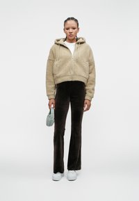Beige fleece zip-up hoodie with drawstring, paired with dark brown flared pants. Model holds a small light blue bag and wears white sneakers.