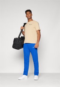 Beige bomull T-skjorte med logo, blå joggebukser og hvite sneakers. Svart treningsbag med mesh-detaljer. Enkel, uformell treningstøy.