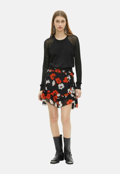 Giovane donna che indossa una maglia nera con maniche in rete, una gonna nera con stampa floreale rossa e bianca, stivali neri fino a metà polpaccio e grandi orecchini floreali.