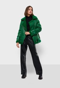Zelená bunda z ovčieho rúna s podšívkou, na zips, s veľkým golierom a detailmi s prackami, skombinovaná s čiernymi koženými nohavicami a čiernymi čižmami.
