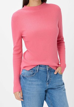 Personne portant un pull rose côtelé ajusté à manches longues, rentré dans un jean en denim bleu clair, la main dans la poche.