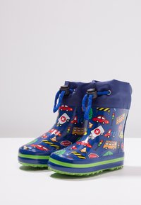 Des bottes en caoutchouc bleues présentent un motif coloré de véhicules et de panneaux de circulation, avec une semelle verte et des lacets bleus pour un maintien sécurisé.