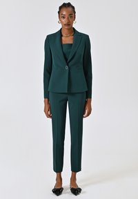 Blazer verde scuro e pantaloni coordinati, taglio sagomato, texture liscia, chiusura con un solo bottone, abbinati a ballerine nere a punta.