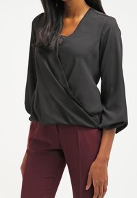 Blusa nera incrociata con scollo a V, realizzata in tessuto liscio, con maniche a 3/4 e sottili dettagli in pizzo sullo scollo. Abbinata a pantaloni bordeaux.