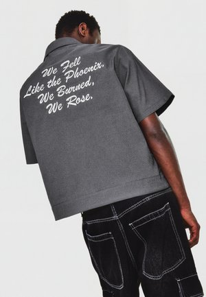 Mann trägt ein graues Kurzarmshirt mit weißem Text „We Fell Like the Phoenix, We Burned, We Rose“ auf dem Rücken und schwarze Hosen mit weißen Nähten.