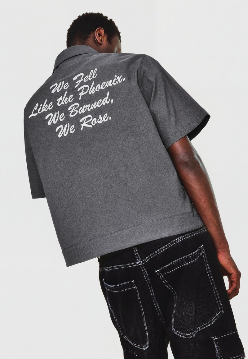 Homme portant un t-shirt gris à manches courtes avec un texte blanc « We Fell Like the Phoenix, We Burned, We Rose » dans le dos et un pantalon noir avec des coutures blanches.