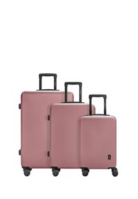 Trois valises rigides roses de tailles variées, avec des poignées rétractables noires et des roulettes. Texture lisse et design minimaliste.