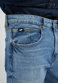 I jeans in denim azzurro chiaro presentano un classico design a cinque tasche, cuciture marroni a contrasto e una piccola etichetta nera del marchio sulla tasca posteriore.