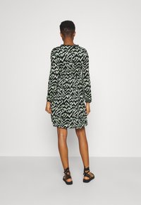 Langärmliges Kleid in grünem und schwarzem abstraktem Muster, mit tailliertem Schnitt und ausgestelltem Rock. Das Material wirkt strukturiert. Trägt schwarze Sandalen.