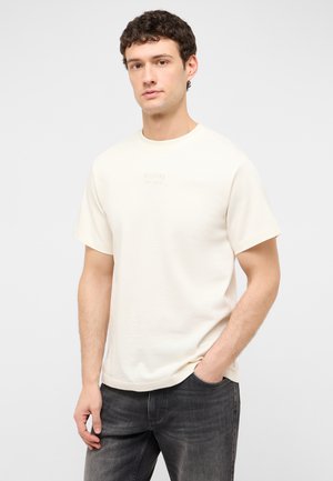Junger Mann mit kurzen lockigen Haaren, der ein schlichtes cremeweißes T-Shirt mit kurzen Ärmeln und dunkelgraue Jeans trägt, vor einem hellen Hintergrund.