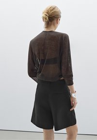 Femme aux cheveux blonds attachés en chignon, portant un haut noir transparent à manches longues, un short noir taille haute et un bracelet argenté, debout contre un mur blanc.