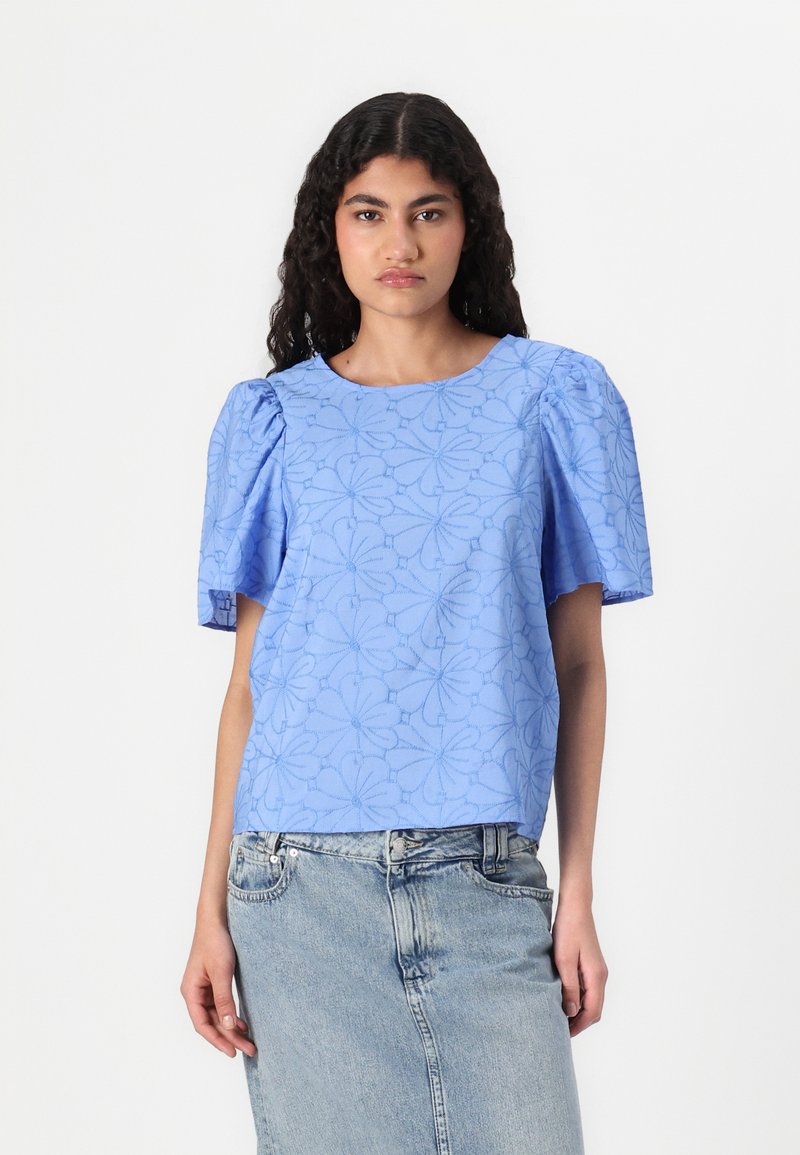 Haut bleu ciel à manches bouffantes avec texture florale, encolure ronde et coupe décontractée. Associé à une jupe en denim claire.