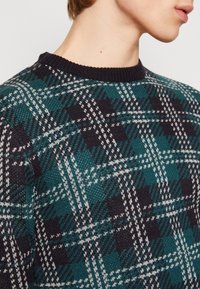Sweter z okrągłym dekoltem w ciemnoturkusowo-czarną kratę z białymi akcentami. Dzianina o fakturowanej strukturze z prążkowanym kołnierzem.