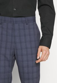Navy plaid trousers made of a textured fabric with a checked pattern, featuring a flat front and side pockets, worn with a black shirt.