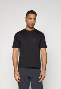 Black short-sleeve t-shirt made of smooth fabric, featuring a small chest pocket and a minimalist design. Worn with dark trousers.
