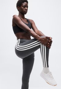 Αθλητικός/ή φοράει μαύρο αθλητικό σουτιέν, γκρι κολάν με λευκές ρίγες και λευκά αθλητικά παπούτσια, τεντώνεται κρατώντας ένα γόνατο σηκωμένο.