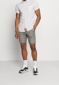 Camicia a maniche corte con bottoni di colore grigio chiaro a righe, abbinata a pantaloni corti a quadretti grigi. Sneakers bianco e nere con calzini bianchi.