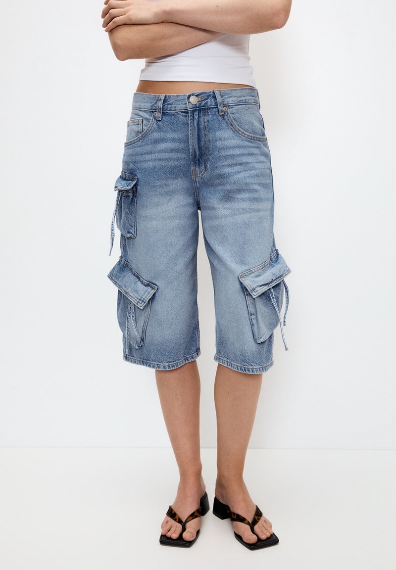 Pantalones cortos de carga azul claro hechos de mezclilla, con múltiples bolsillos laterales y un corte relajado, de longitud hasta la rodilla con dobladillo deshilachado y un ligero desgaste.