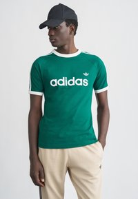 Πράσινο κοντομάνικο T-shirt με λευκή επένδυση και λογότυπο "adidas" σε έντονα γράμματα. Παρουσιάζεται με μαύρο καπέλο και μπεζ παντελόνι.
