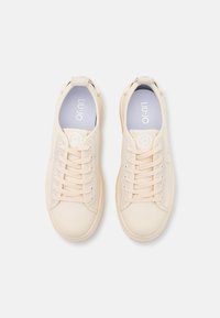 Un paio di sneaker beige stringate con solette grigio chiaro, viste dall'alto su uno sfondo bianco.
