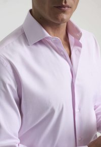 Camisa de dress de manga larga color rosa claro, confeccionada en una tela suave con un cuello clásico y botones blancos, que presenta un logo sutil en el pecho.