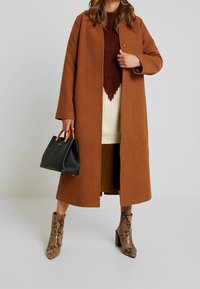 Ruskea villasekoitteen päällystakki suoran muotoilun kanssa, yhdistettynä viininpunaisiin neulepaitoihin ja mustaan käsilaukkuun. Teksturoitu käärmeennahkasaapas nilkkoihin.