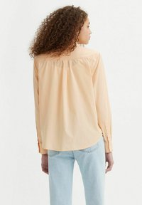 Lichtoranje langetermijn shirt met knoopmanchetten, een geplooide rugplooi en een ronde zoom, gecombineerd met lichtblauwe jeans.