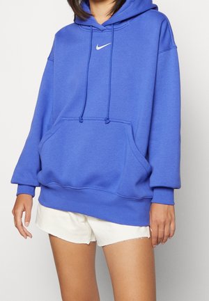 Vrouw draagt een blauwe Nike hoodie met wit logo en voorzak, gecombineerd met witte shorts met rafelige randen.