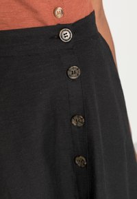 Vêtement noir avec une texture lisse, présentant une rangée de quatre boutons à motifs sombres sur le côté. Gros plan sur le tissu et les détails des boutons.