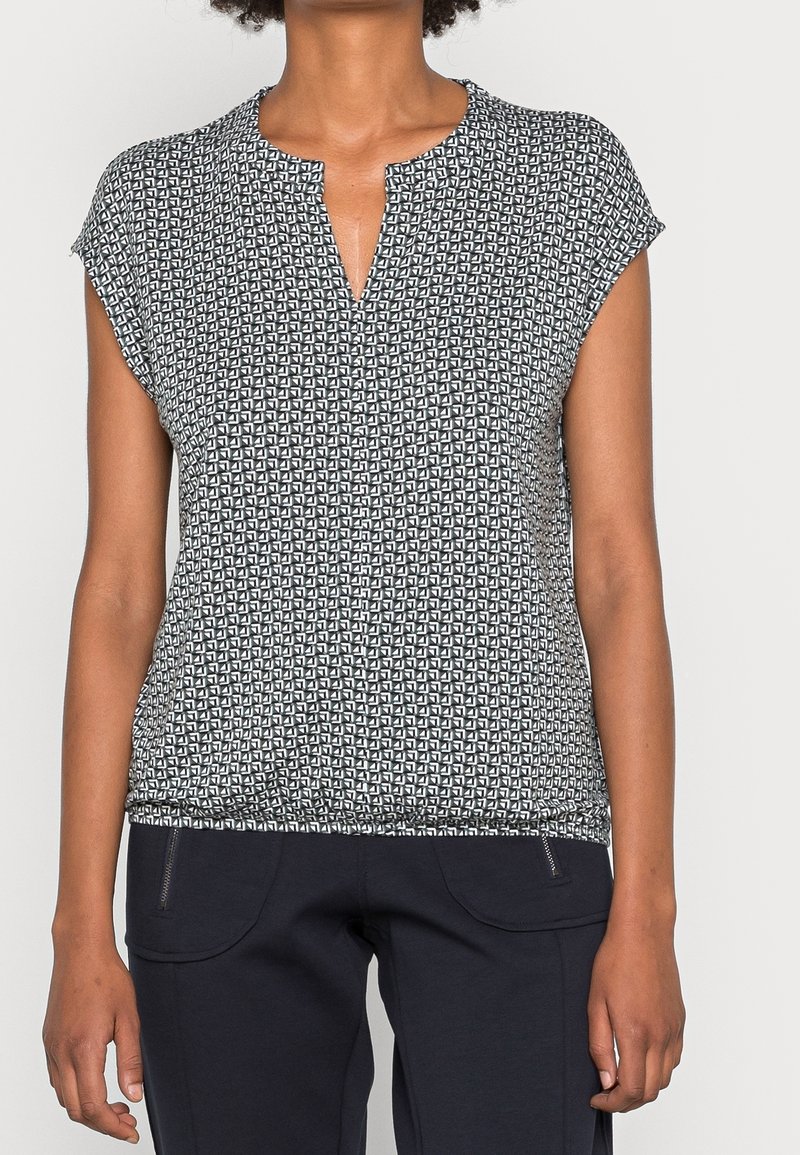 Femme portant un haut à manches courtes à motifs noir et blanc associé à un pantalon noir avec poches zippées à l'avant.