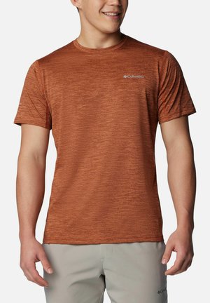 Mann trägt ein rostfarbenes Columbia Kurzarm-T-Shirt und hellgraue Hose, lächelt und steht mit entspannten Händen.